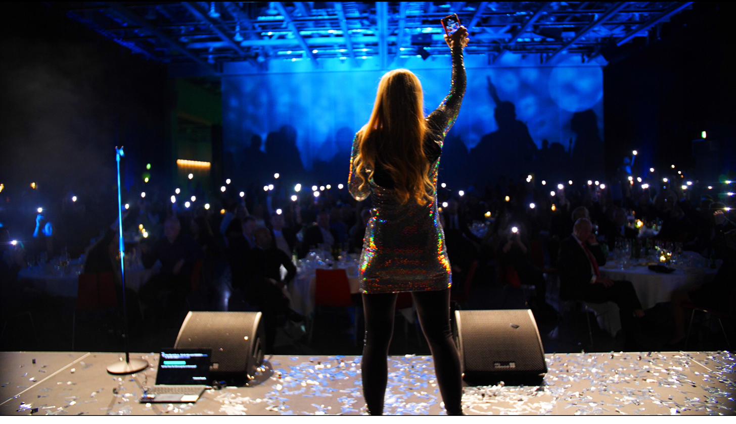 Anna Jois on stage in front of an audience holding up phone lights